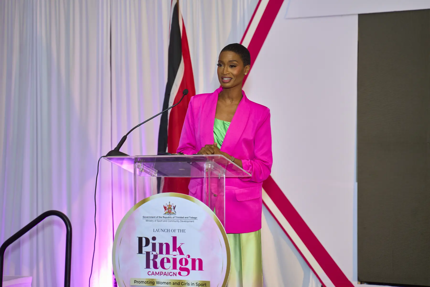 Ainka Will at a podium in a magenta blazer beside the Trinidad and Tobago flag