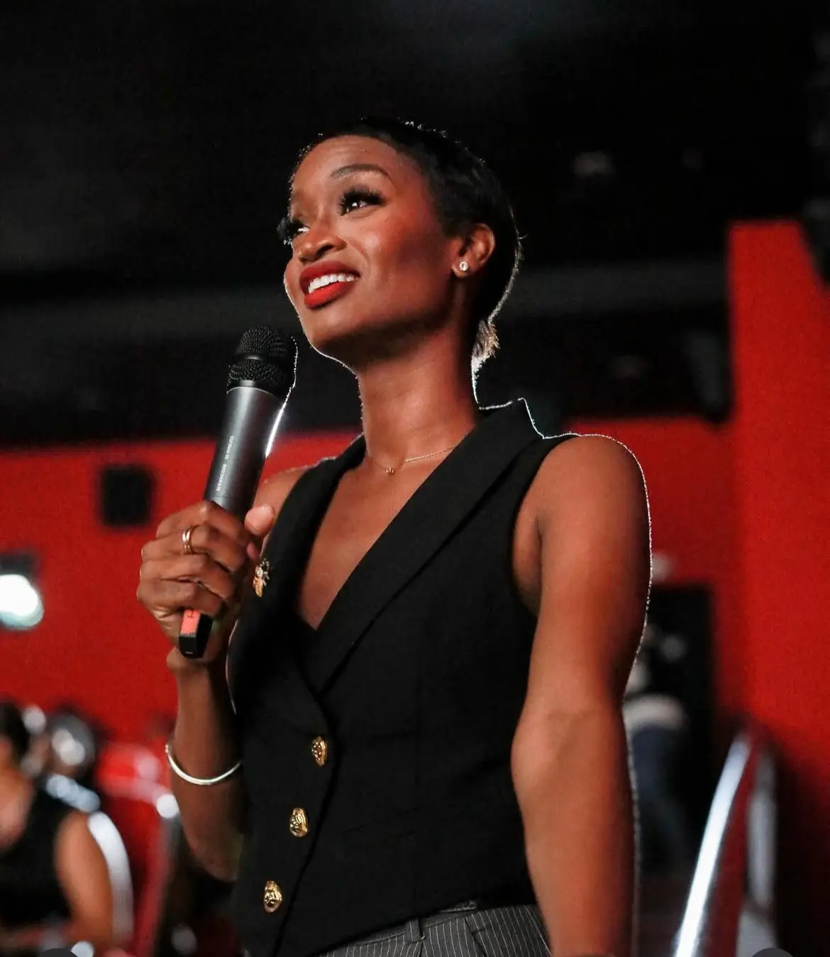 Ainka Will holding a microphone in a black vest against a red backdrop