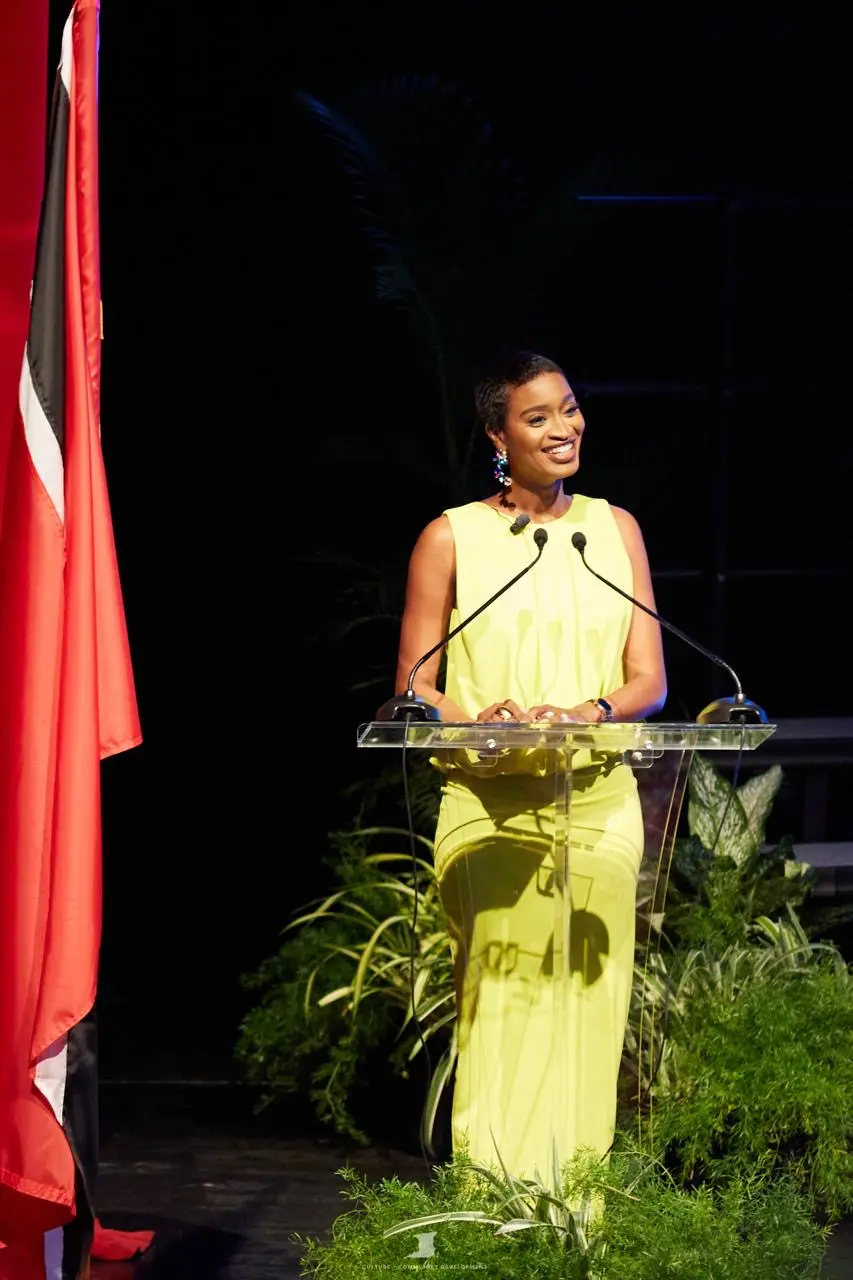 Ainka Will at a podium in a lime dress beside the Trinidad and Tobago flag
