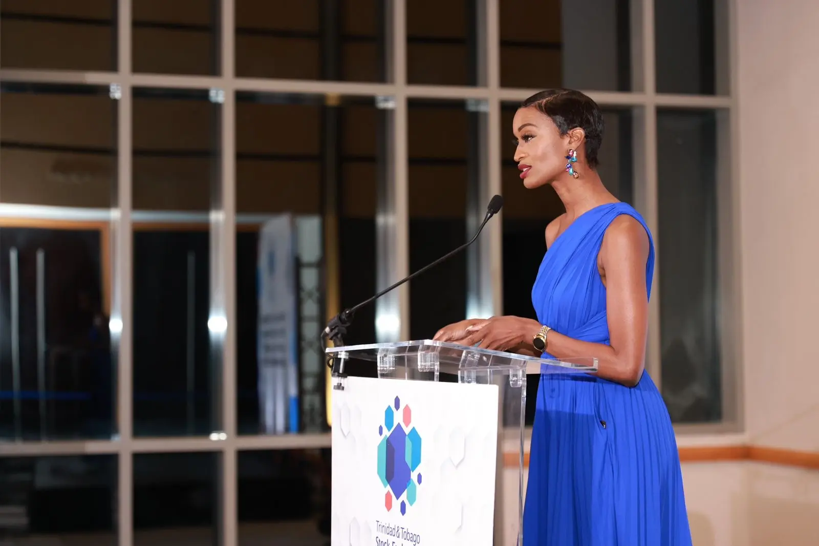 Ainka Will in a blue one-shoulder gown speaking at the Trinidad and Tobago Stock Exchange podium