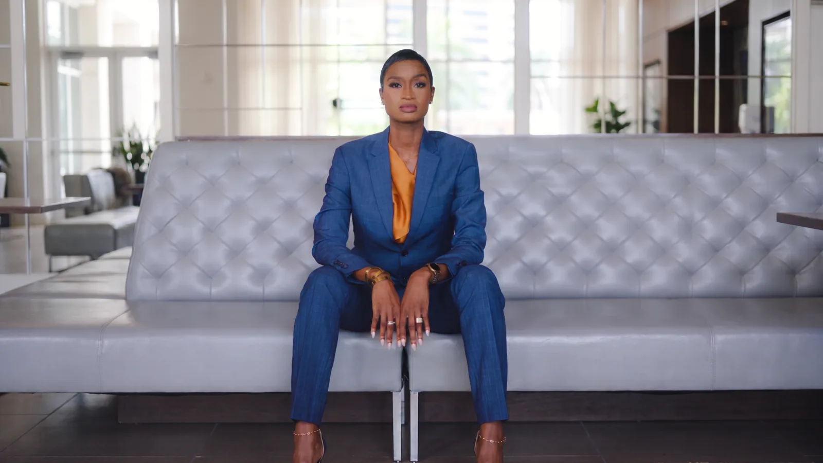 Ainka Will seated in a blue suit and orange silk blouse on a leather banquette