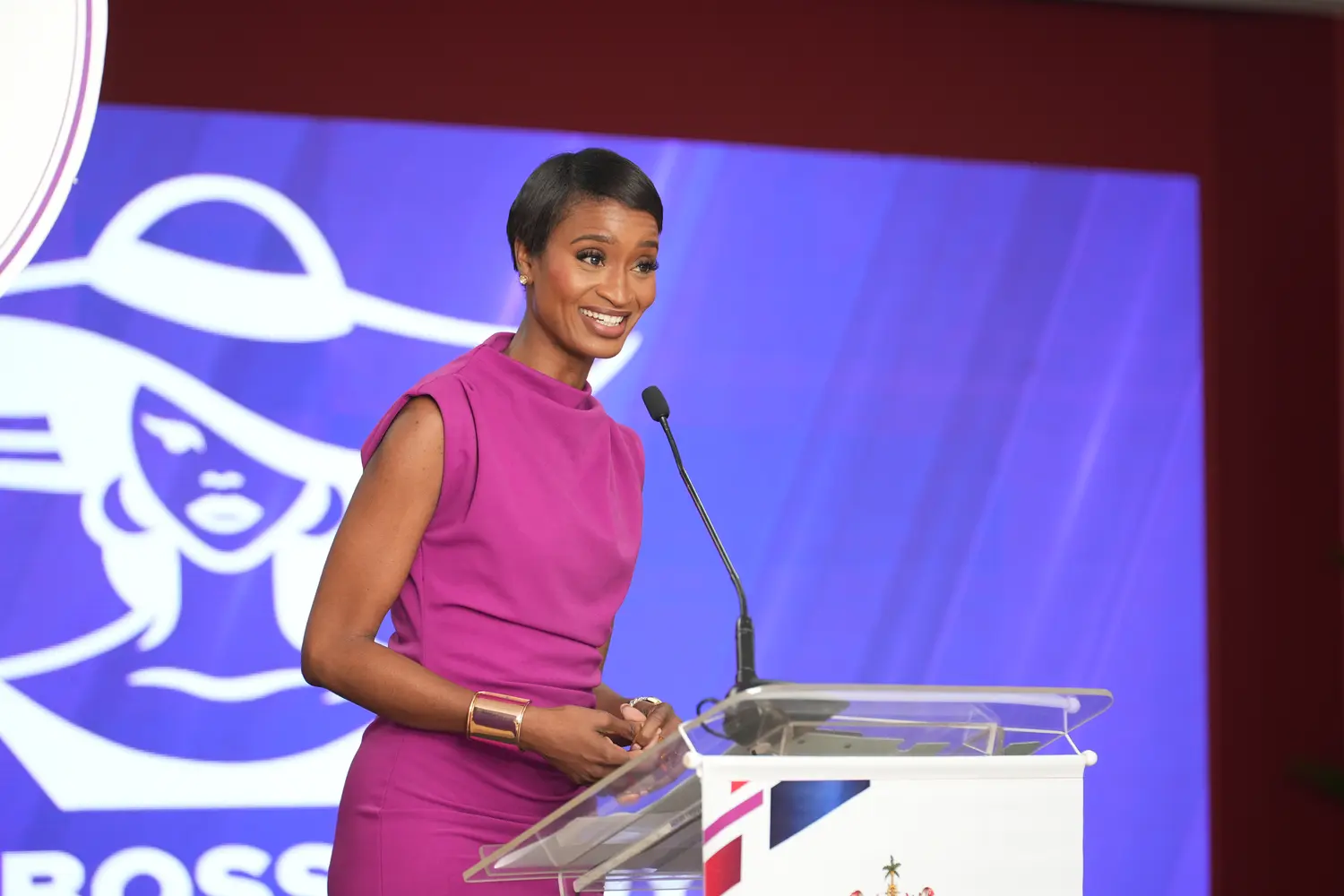 Ainka Will at a podium in a magenta dress addressing an audience