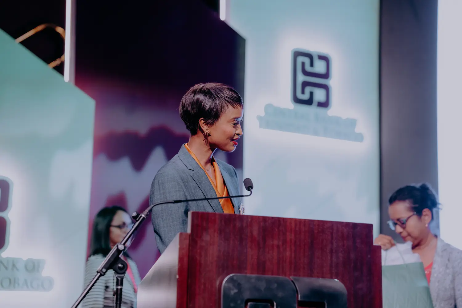 Ainka Will speaking at a lectern in a grey blazer
